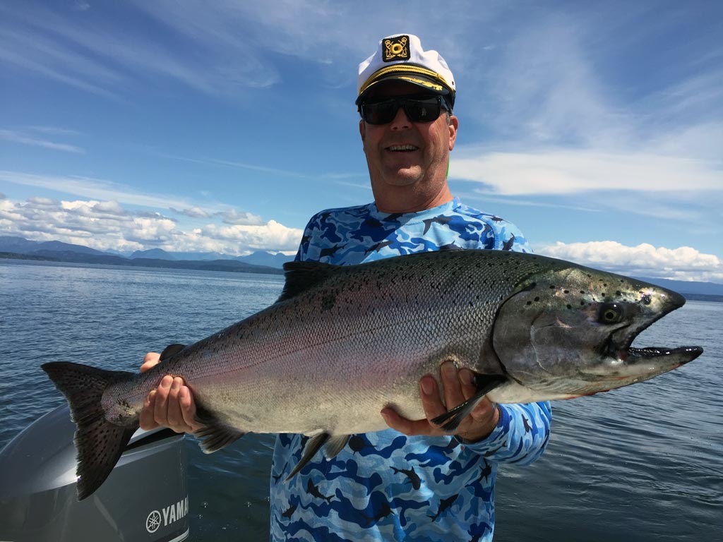 A man with a captain's hat smiling and holding a large salmon.