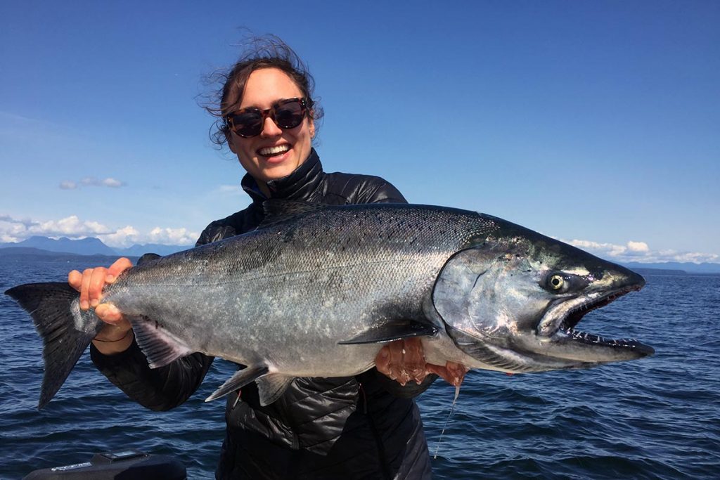 A happy lady holding a large salmon with the ocean in the background.